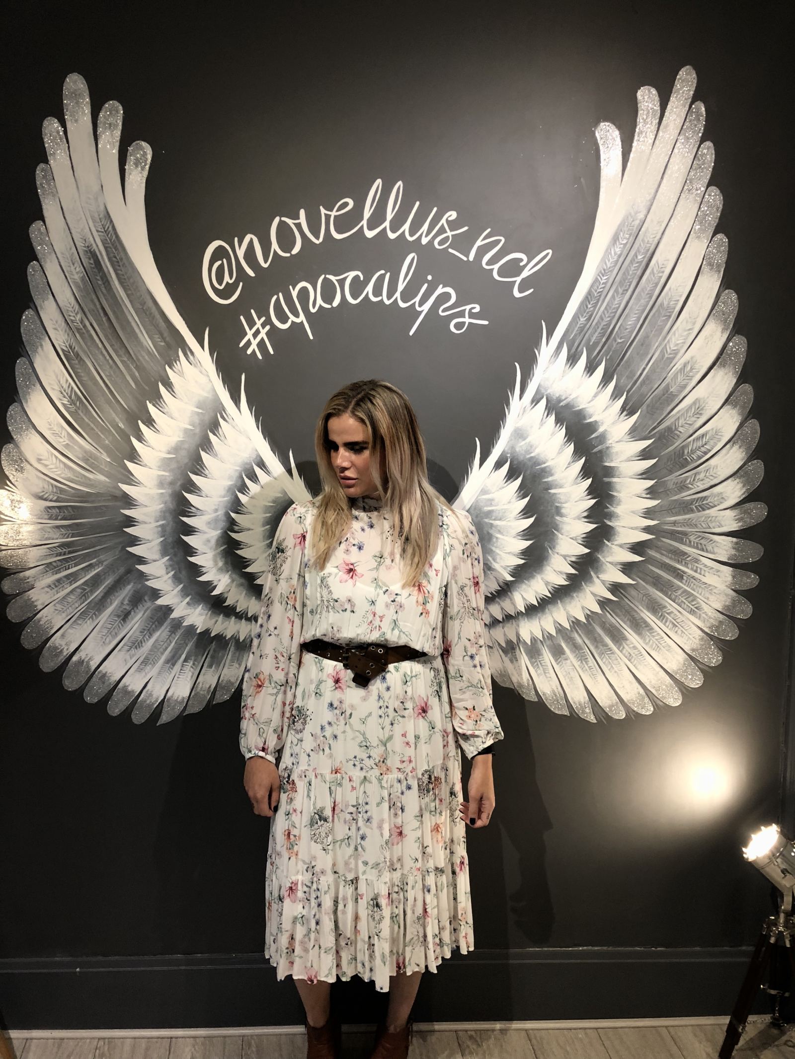 Blogger Pixie Tenenbaum standing in front of Angel wings painted on a dark grey feature wall in the new Novellus Aesthetics clinic room at 1 Benton Terrace in Jesmond. Glitter tipped wings lit up by a cinema type floor lamp featuring the words Novellus newcastle and #Apocalips. taken for Fashion Voyeur blog